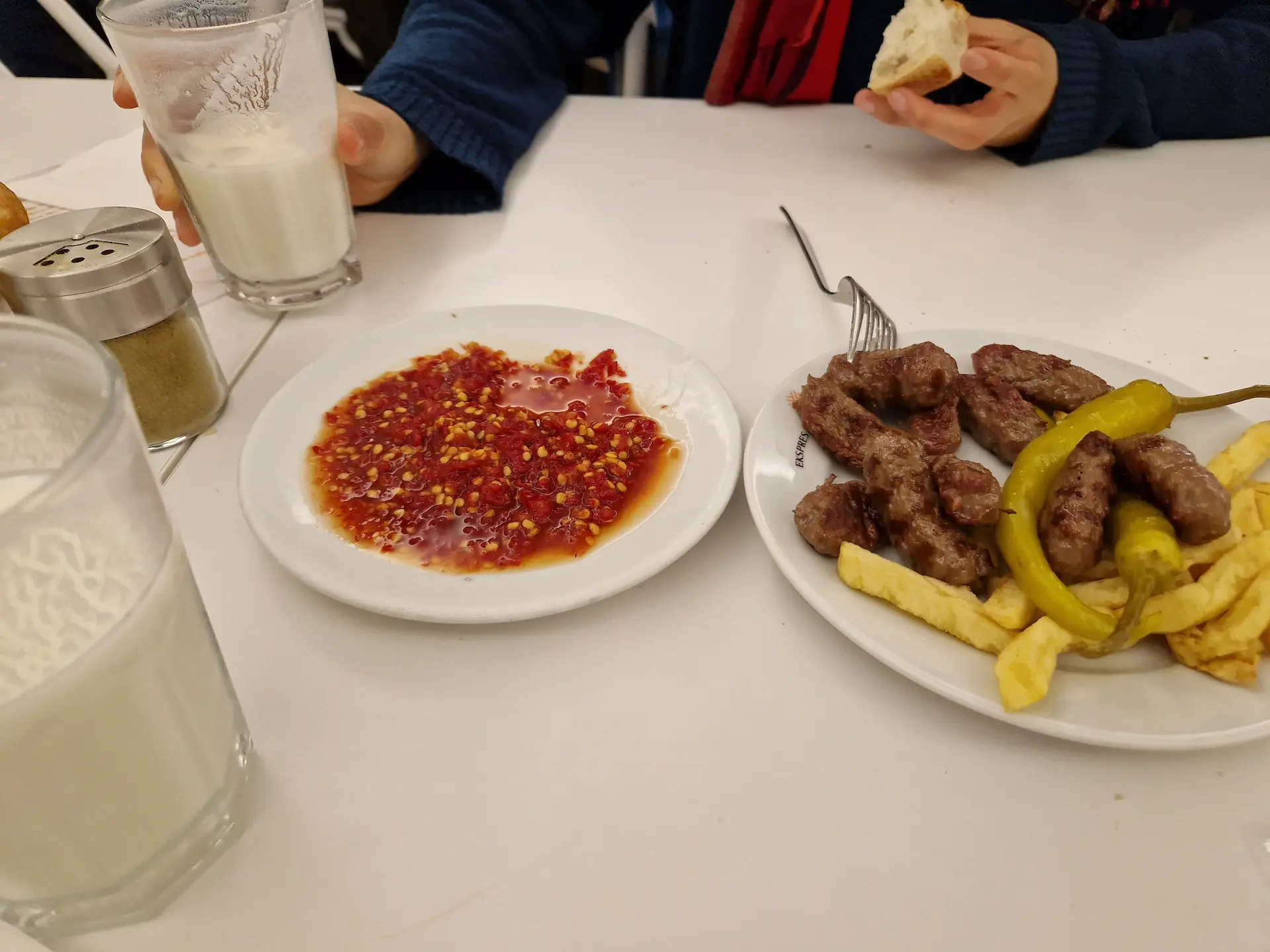 Assiette de İnegöl köfte à Ekspres İnegöl Köftecisi, on voit les boulettes de viande servies avec des frites, et l'assiette entière de sauce au piment rouge à côté. La main de Clara est sur la photo, elle tient un verre de ayran.