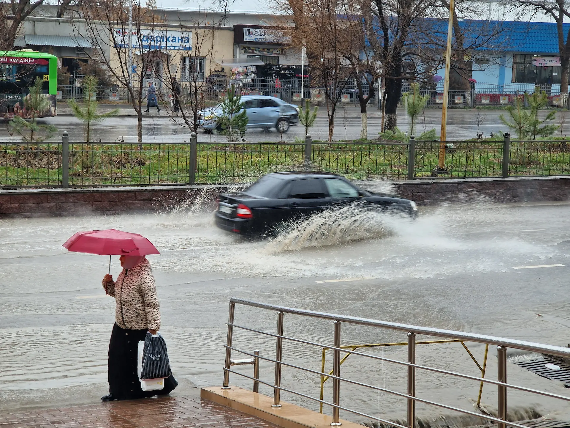 Une dame passe avec un parapluie, une voiture roule dans une grosse flaque, créant une énorme éclaboussure.