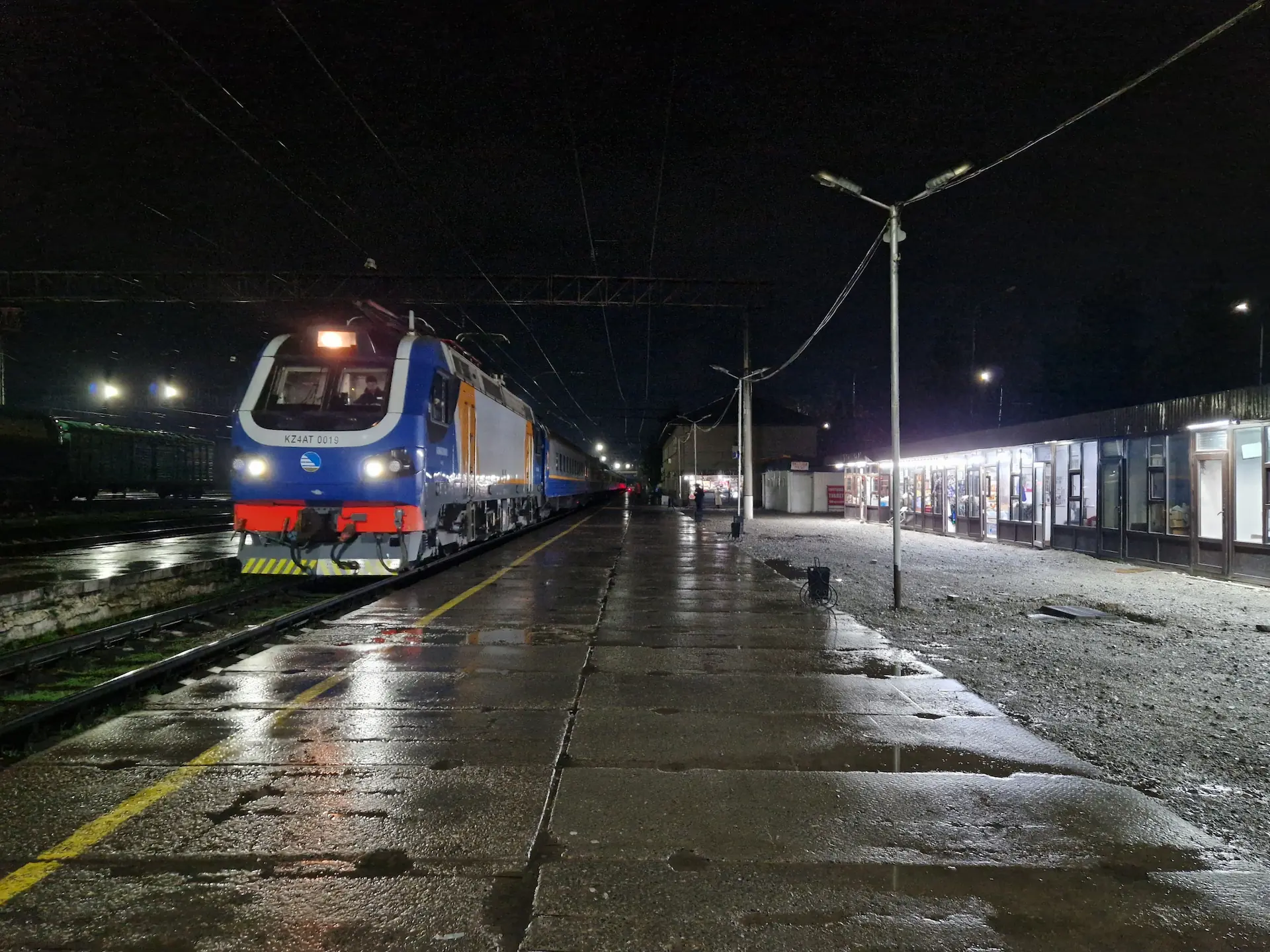 Il fait nuit. Le train arrive à la gare, où le sol est encore mouillé de la pluie.