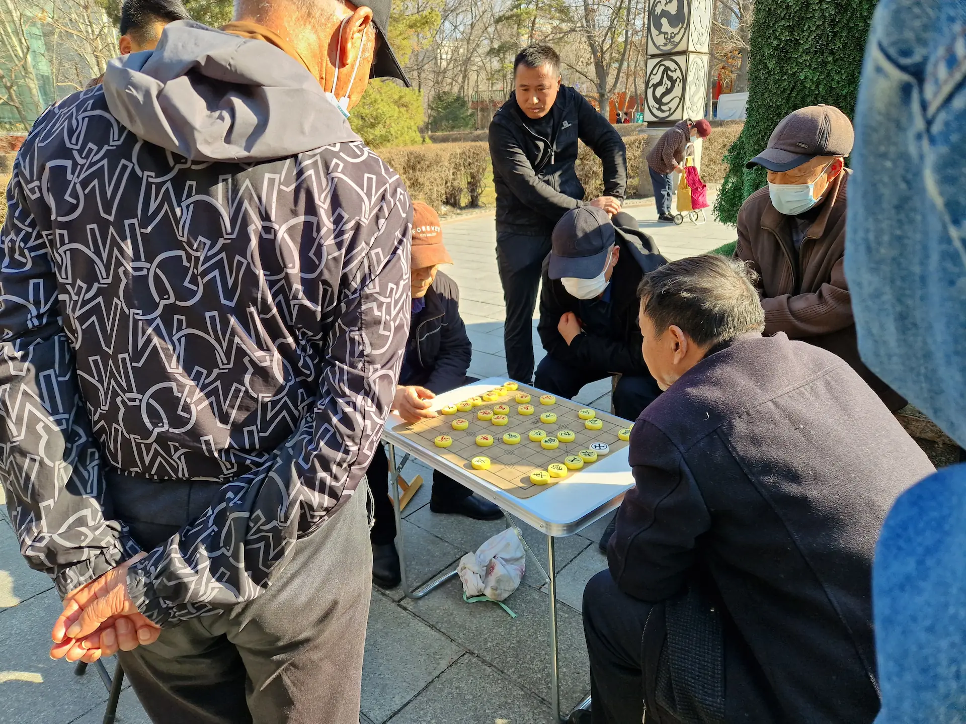 Deux messieurs s'affrontent au xiangqi sur une petite table en plastique pliable. Autour d'eux, six personnes observent le jeu intensément.