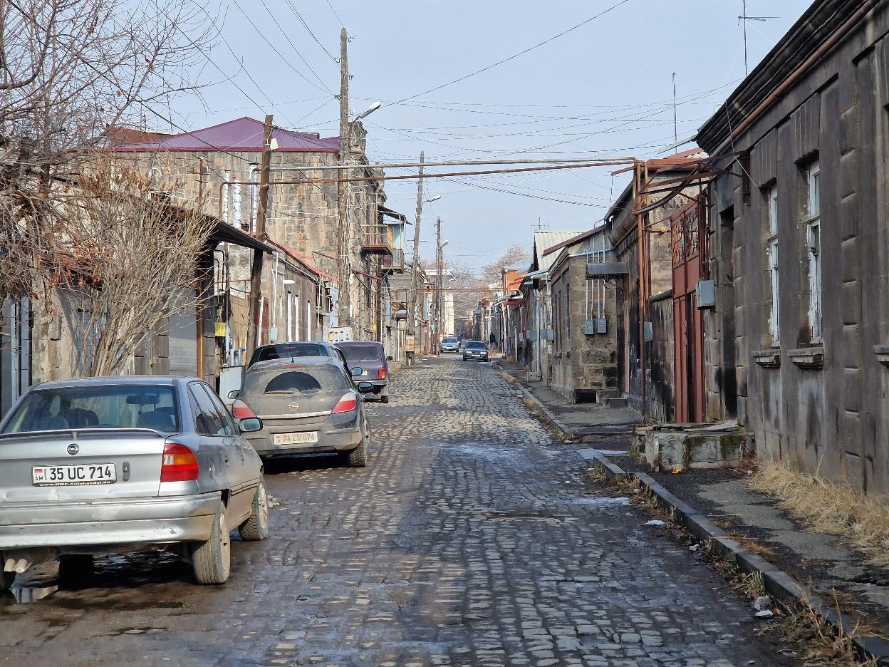 Une rue de Gyumri, de vieux bâtiments gris à un ou deux étages bordent la rue. Le sol est dallé, des vieilles voitures sont garées sur le côté. De la plomberie et des raccordements électriques traversent le ciel au-dessus des bâtiments.