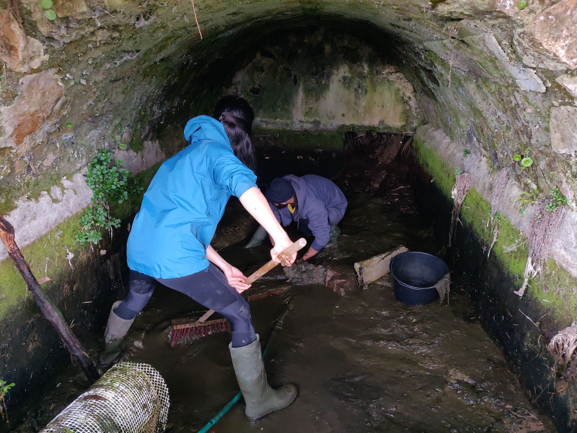 Clara, dans la source maintenant presque vide, utilise un balai de cantonnier pour ramener l'eau qui reste vers l'embouchure du tuyau. Niveau 2.