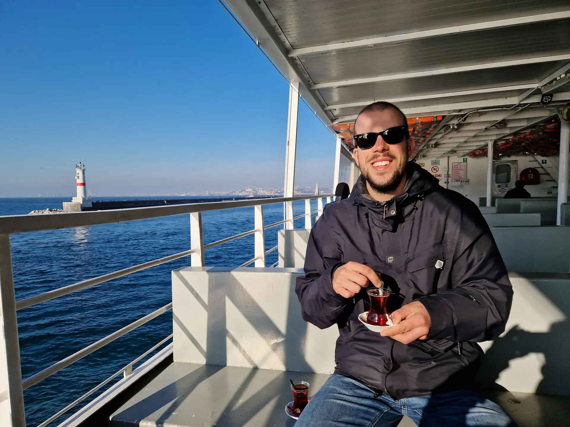 Robin boit un thé sur le pont d'un ferry, il fait grand beau et il porte des lunettes de soleil.