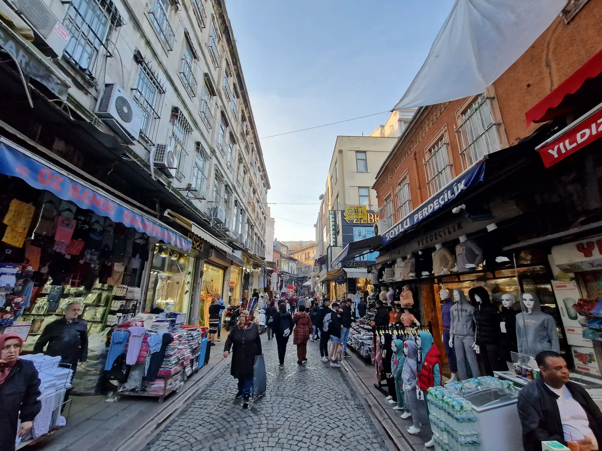 Une ruelle animée d'Eminönü. Sur les deux côtés, des magasins qui vendent des habits, on voit notamment des têtes de mannequins couvertes de voiles colorés. Il y a du monde.