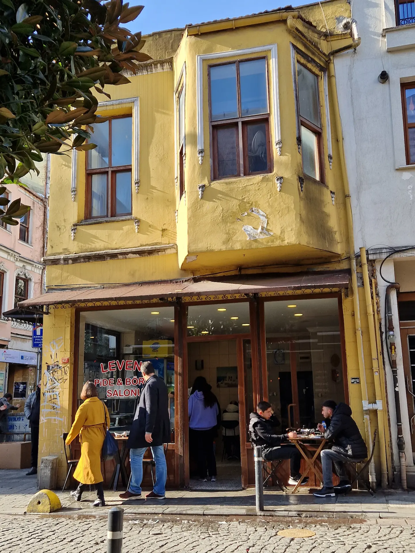 Un petit immeuble de deux étages de Yeldeğirmeni, façade peinte en jaune, des gens assis à une petite table installée devant. Une femme passe avec un imper de la même couleur que la façade.