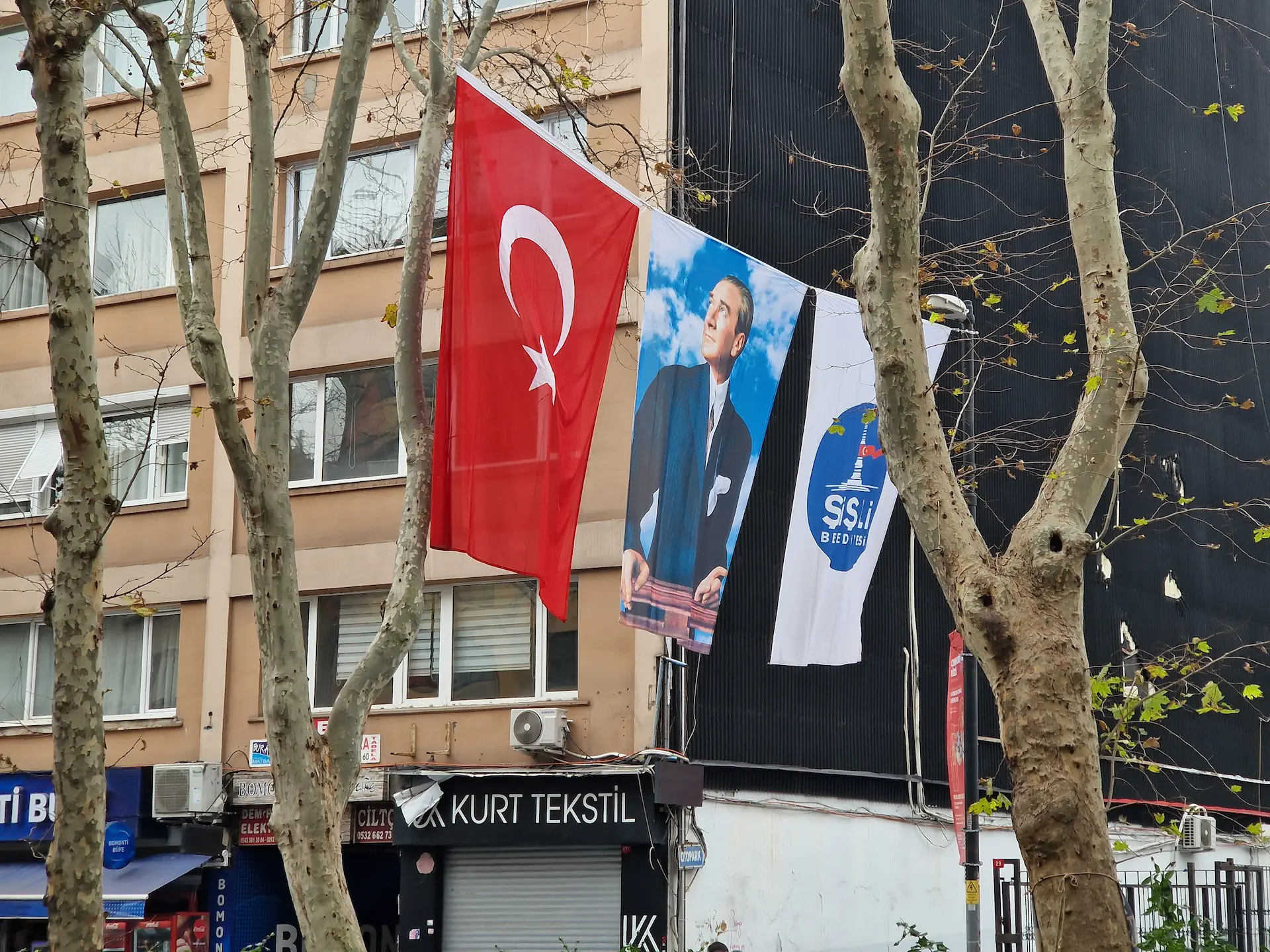 Trois bannières accrochées au-dessus de la route. À gauche : le drapeau turc. Au milieu : Atatürk. À droite : un symbole inconnu.
