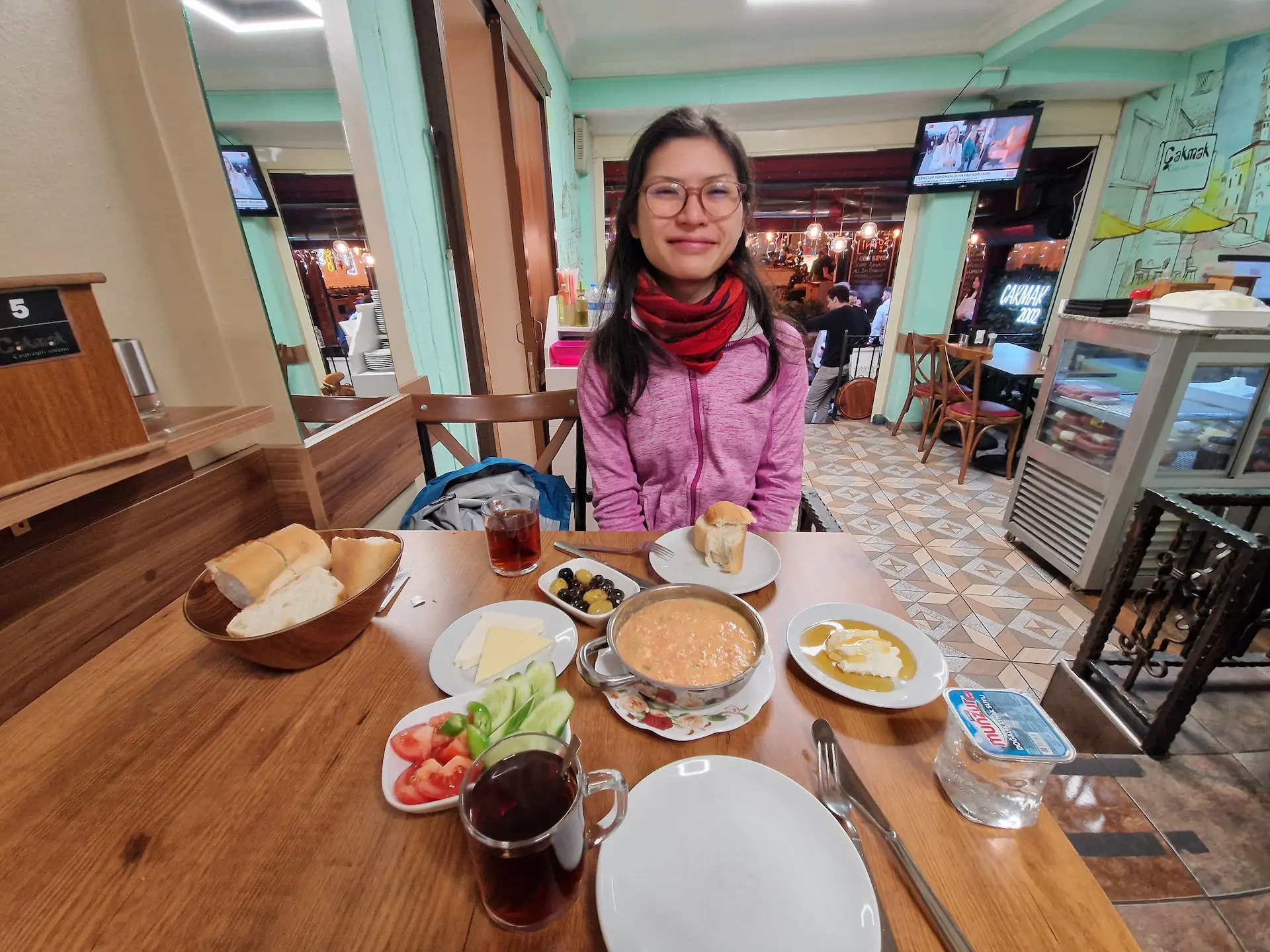 Clara devant un petit-déjeuner turc dans la ruelle du petit-déjeuner, on voit une omelette, des légumes crus, de la crème et miel, du fromage, des olives, du pain. Une télé est allumée en fond, on est presque seuls dans le restaurant.