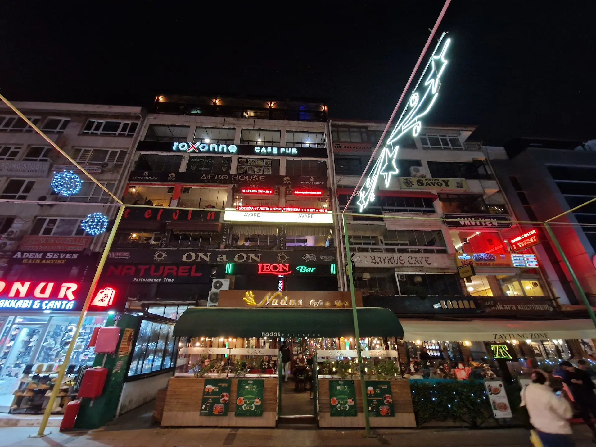 De nuit, un bâtiment illuminé de néons de toutes les couleurs. Les noms de différents magasin, restaurants, cafés, etc. s'entassent sur plusieurs étages.