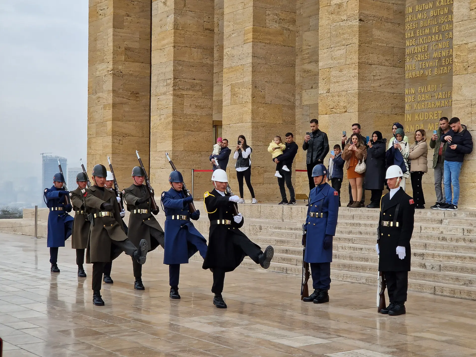 Une parade de neuf gardes passe devant le mausolée. Ils ont des habits militaires cérémonieux, des casques et un fusil chacun. En fond, les visiteurs les filment et les prennent en photo.