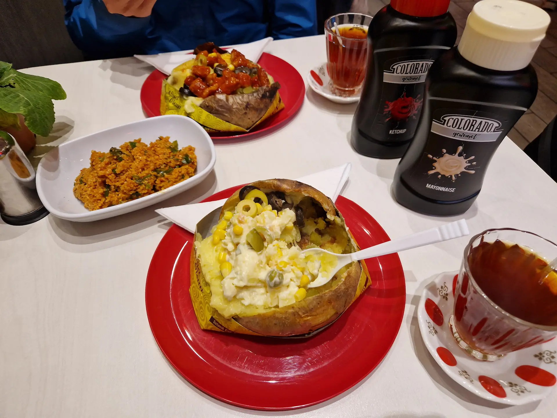 Sur une table, deux plats de kumpir aux ingrédients différents. Entre les deux plats, une entrée de couscous, deux verres de thé. Sur le côté, deux grands pots de ketchup et mayonnaise.
