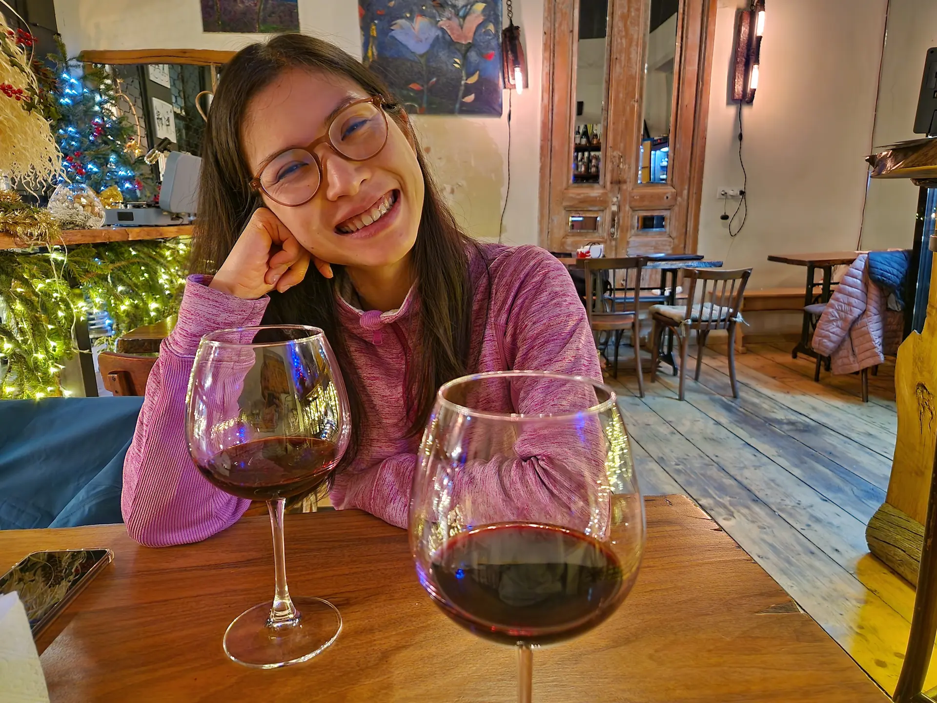 Deux verres de rouge sur une table, Clara assise en face sourit à l'objectif.