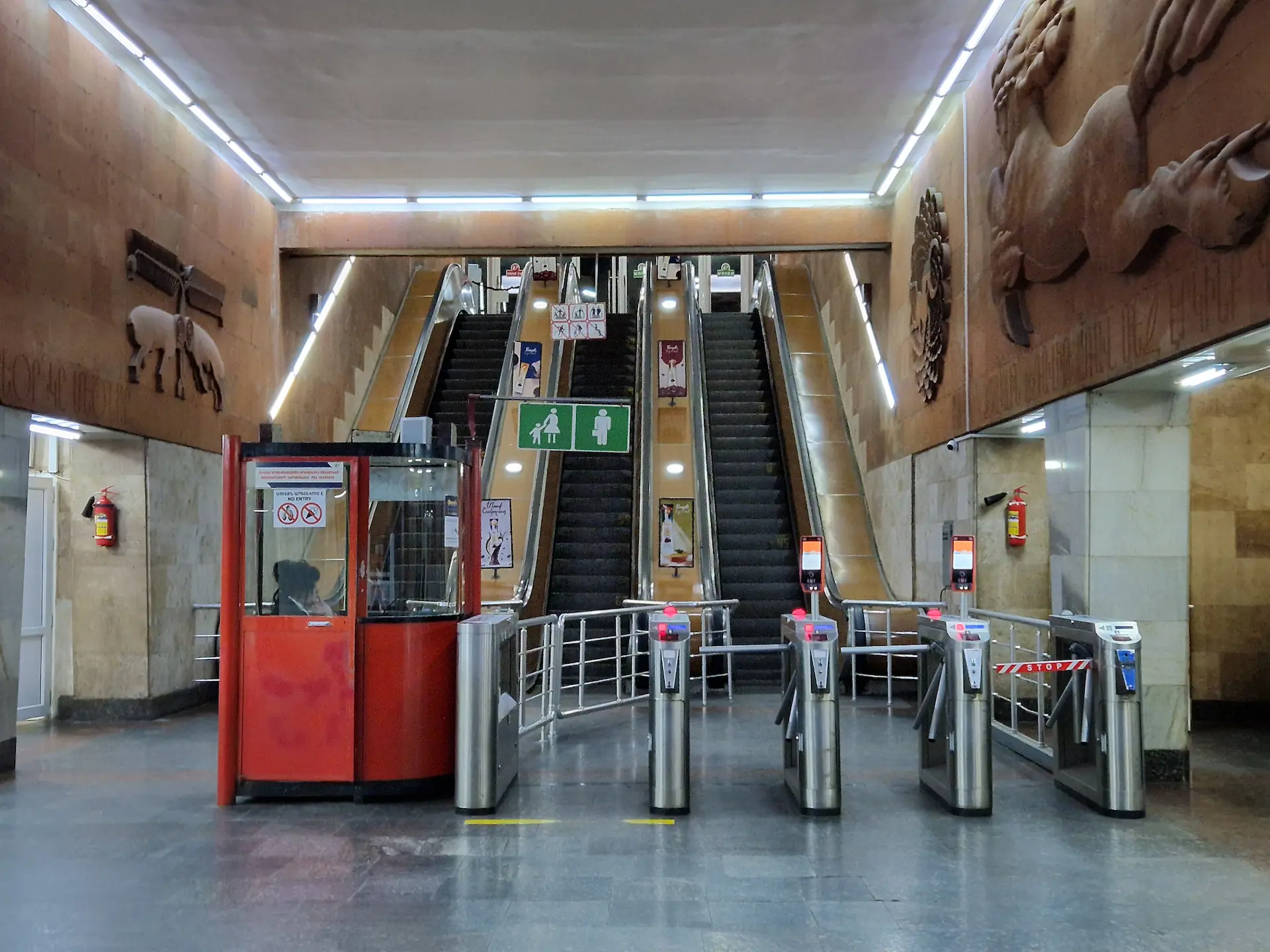 Entrée de la station de la gare. Il y a des fresques soviétiques sur les murs. Devant nous, une cabine rouge où se trouve une employée, trois tourniquets qui mènent à des escaliers roulants.