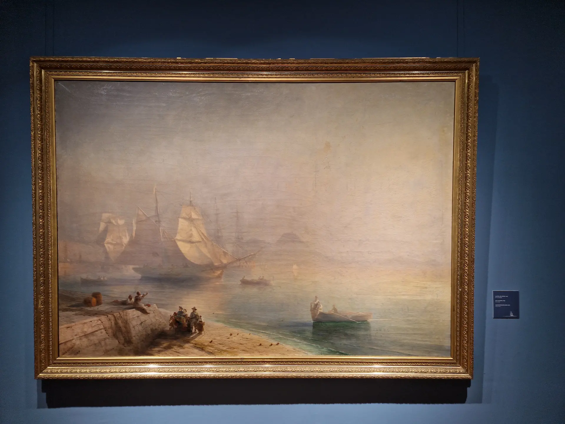 Peinture réaliste de la mer vue du port de nice, il y a du brouillard, on distingue des bateaux. Il y a quelques personnes sur le quai.