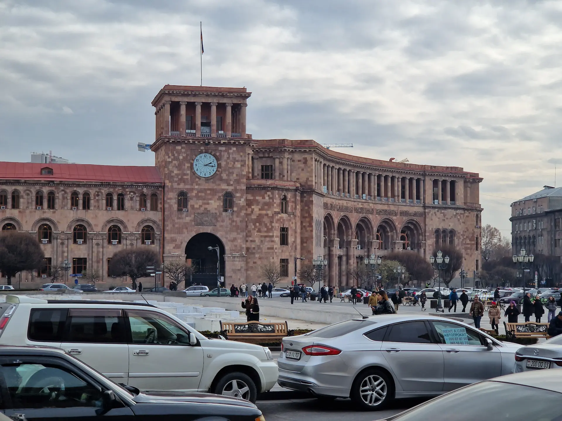 Un grand immeuble de pierre rose à la place de la République à Yerevan, avec du trafic, du monde, une horloge sur la façade.