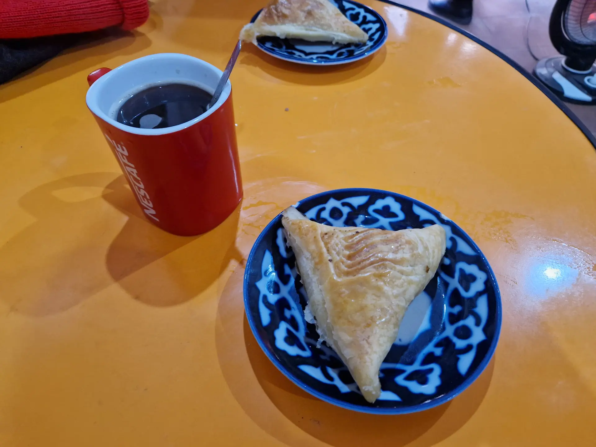 Samsa triangulaire et café noir instantané dans une tasse Nescafé rouge, sur une petite table en plastique orange.