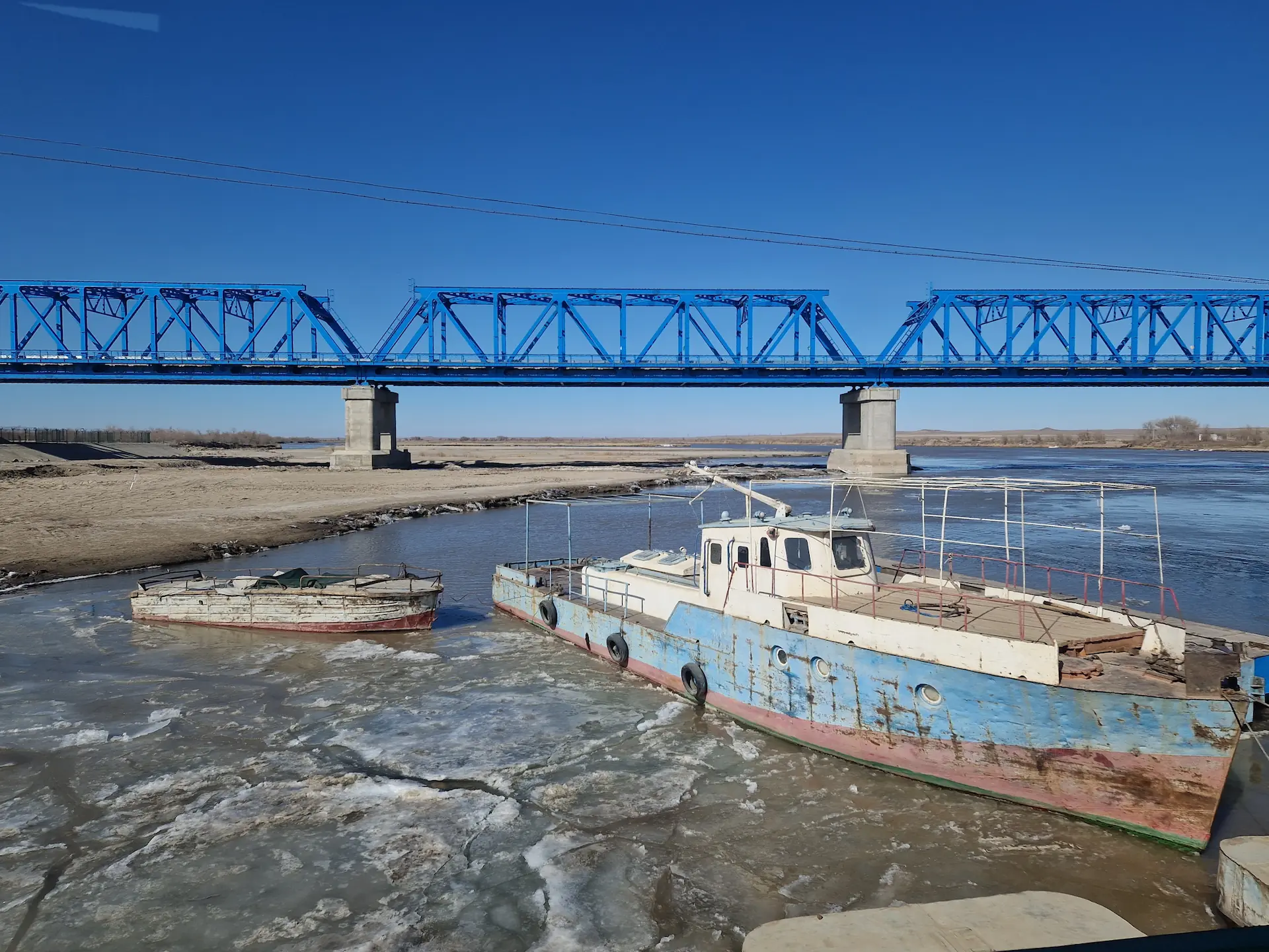 On traverse le pont de l'Amou-Darya dont les morceaux gelés s'éloignent en flottant aux côtés d'un bateau. En fond, un autre pont bleu.