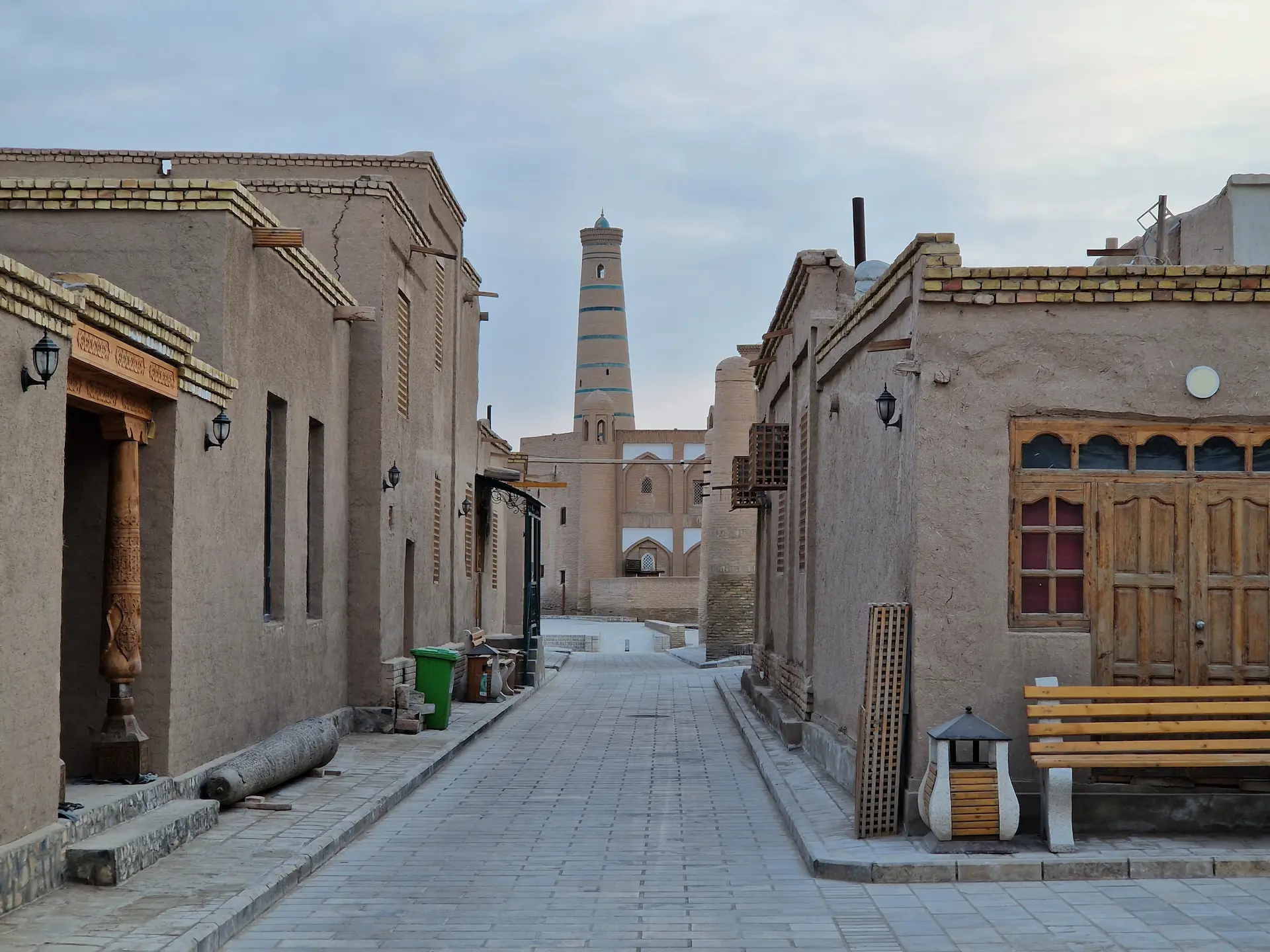 Rue bordée de maisons basses en terre-paille et aux fenêtres en bois. Au bout de la rue, on aperçoit un minaret.