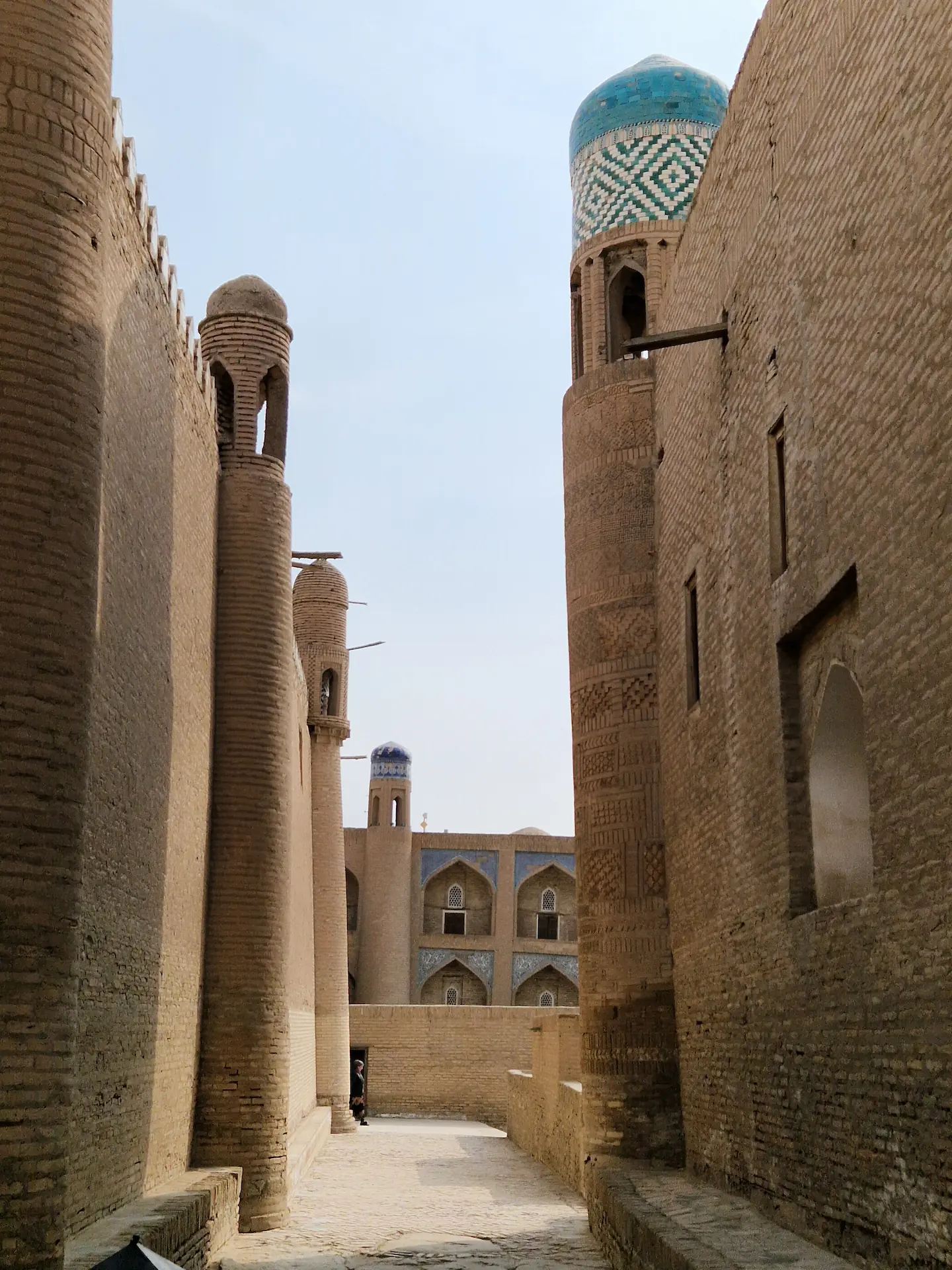 Une petite rue vide entre deux grands murs en pierre. On voit une madrassa en fond.