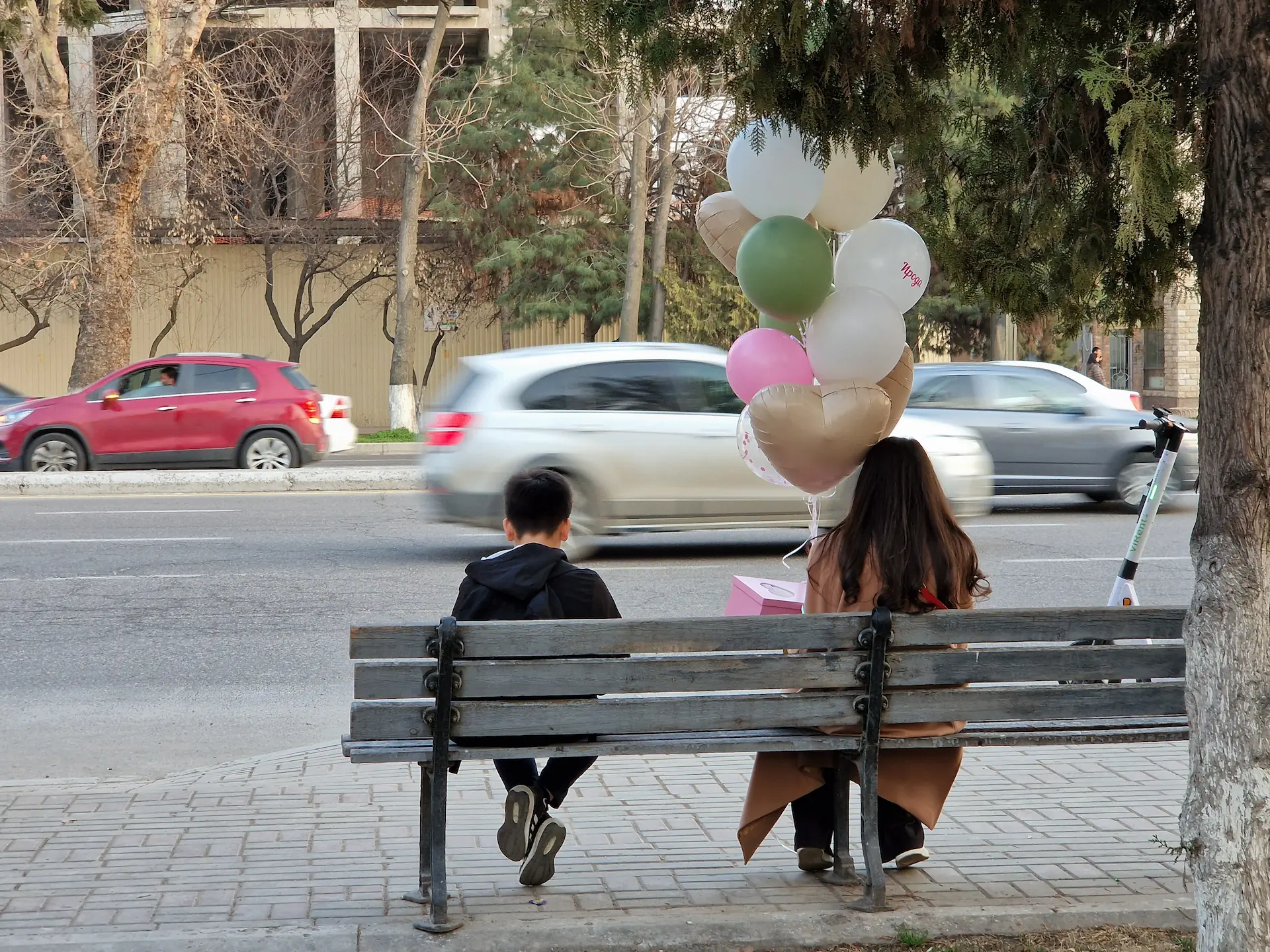 Une femme de dos sur un banc, avec en main un paquet cadeau et une dizaine de ballons d'hélium