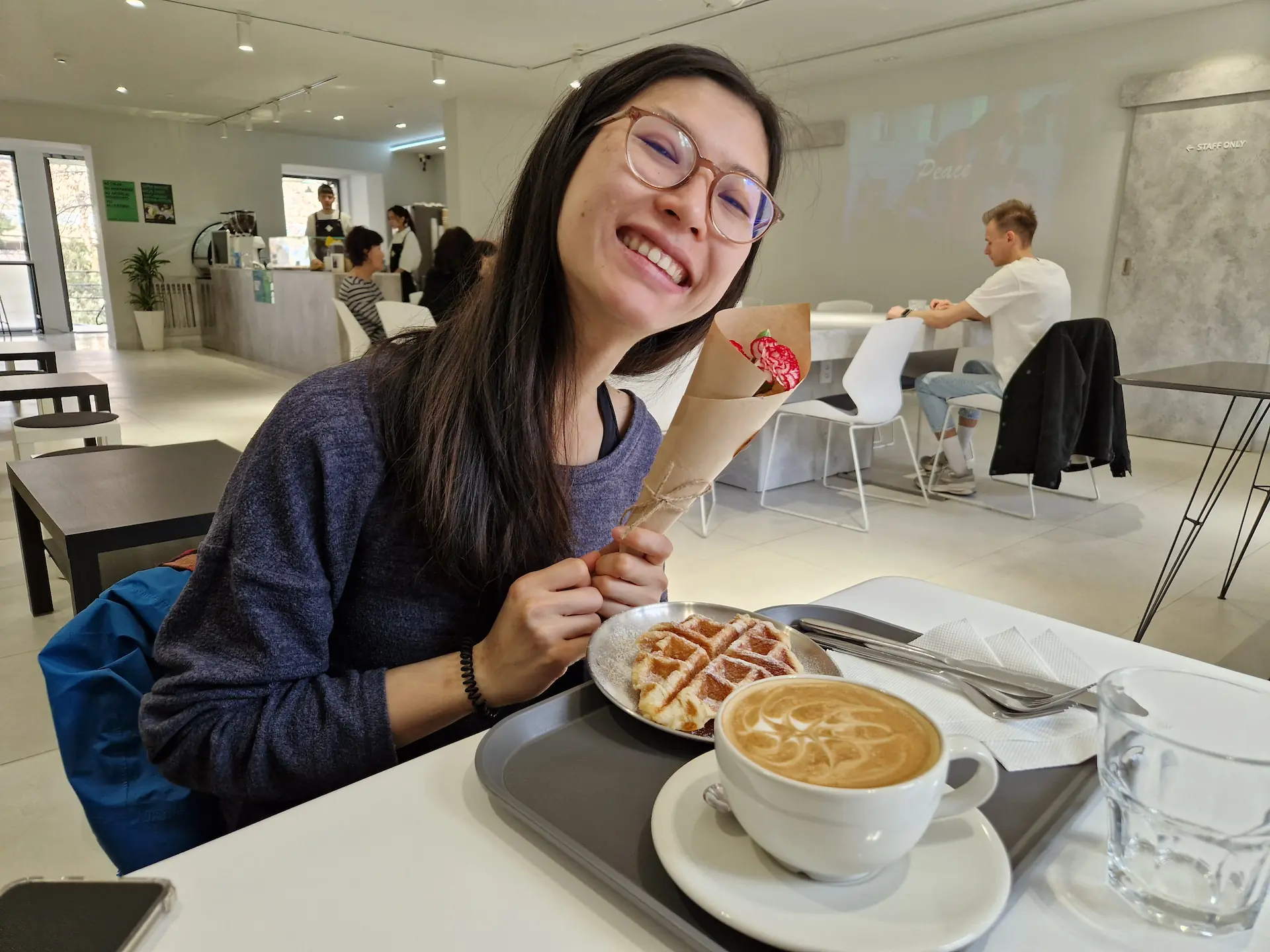 Clara avec son cappuccino, sa gauffre, et une petite fleur dans un café tout décoré de blanc