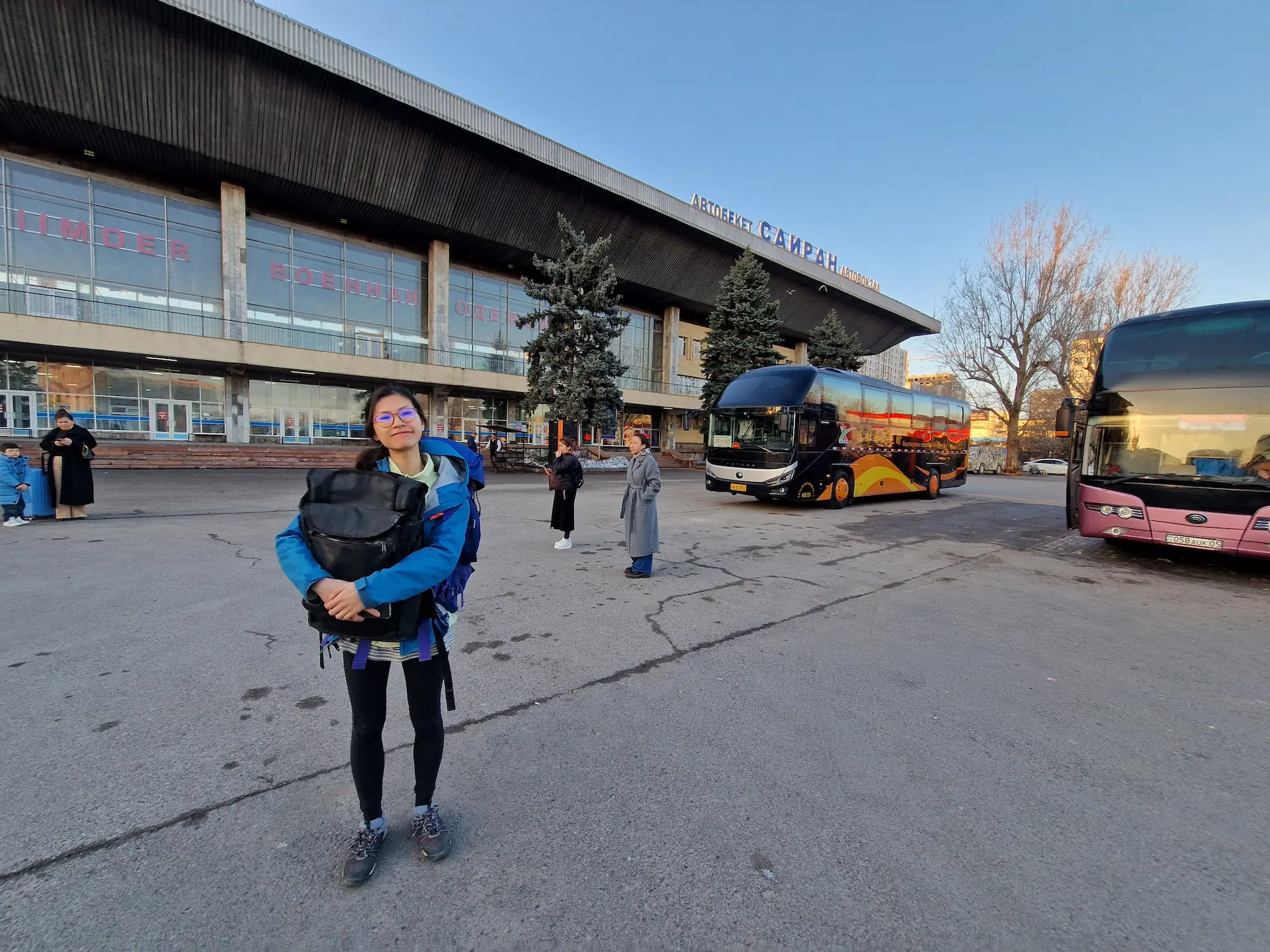 Clara avec son sac à dos à la station de bus d'Almaty au lever de soleil, devant deux grands cars