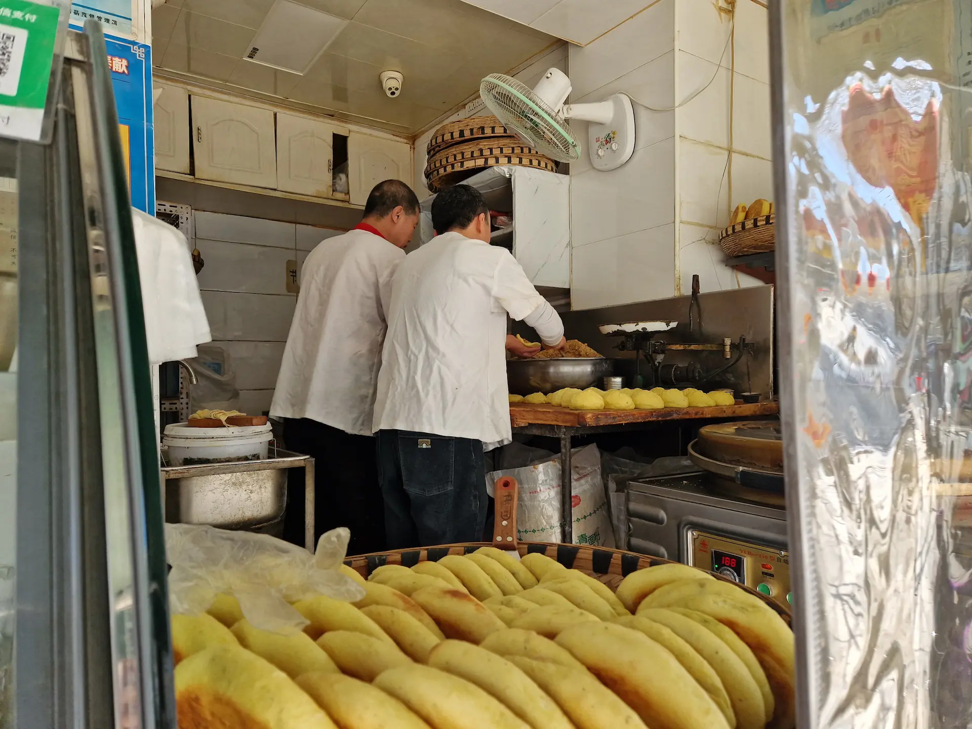 Deux hommes de dos dans une petite boulangerie préparent des bǐng, petits pains plats fourrés qu'on voit à l'avant de la photo