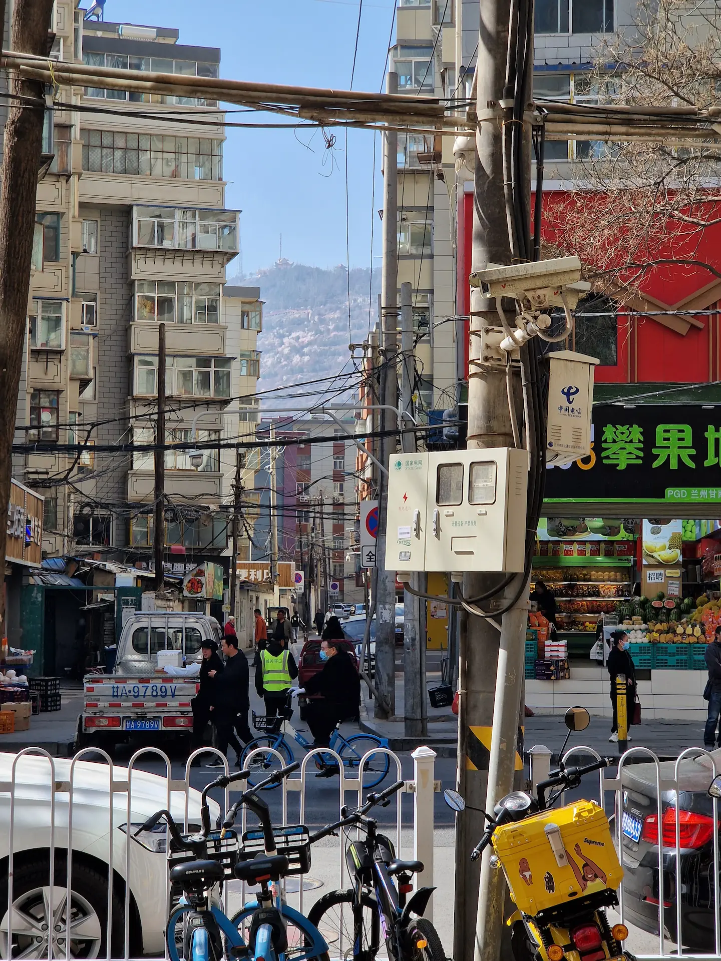 Vue d'une petite rue chaotique, petits bâtiments, vélos, fils électriques, piétons, magasins, une montagne en fond