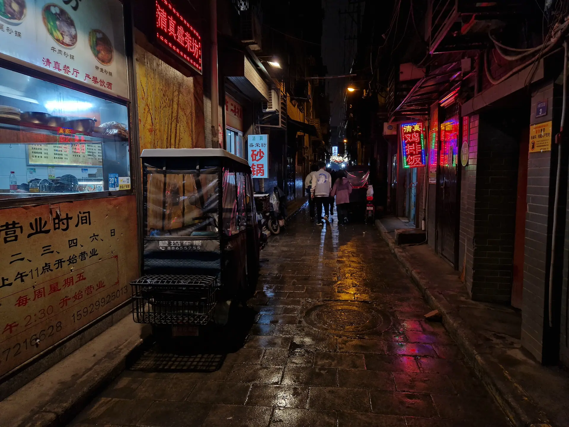 Une ruelle sombre presque déserte après la pluie, où les signes colorés se reflètent sur les pavés mouillés