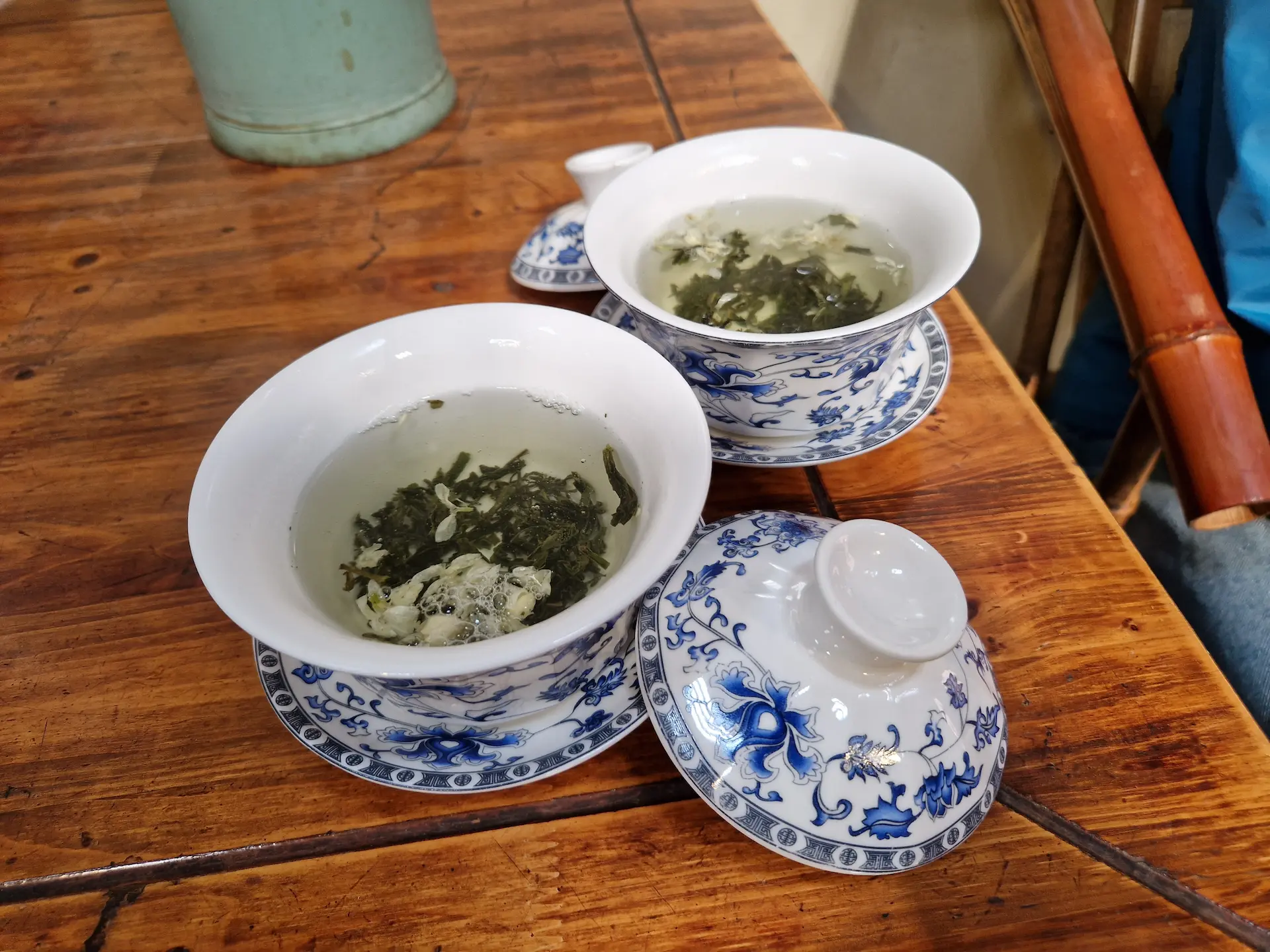 Deux tasses de thé en céramique bleues et blanches. Les feuilles de thé et fleurs de jasmin flottent à la surface de l'eau. Leurs couvercles sont posés sur le côté.