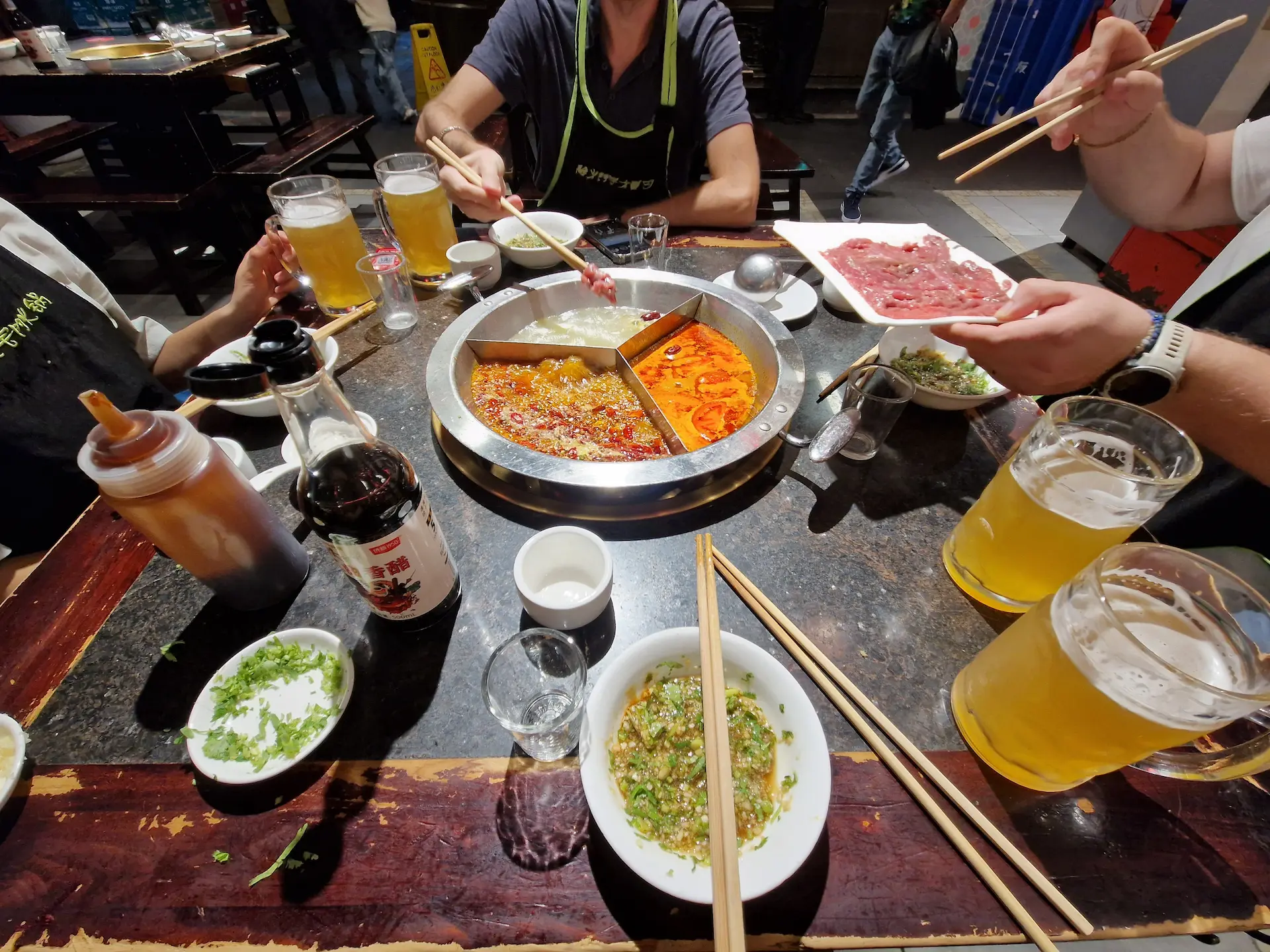 Au milieu de la table, le hotpot avec trois bouillons différents. On est quatre autour de la table, chacun avec sa grande bière fraîche. La personne sur la droite s'apprête à mettre à cuire des morceaux de viande.