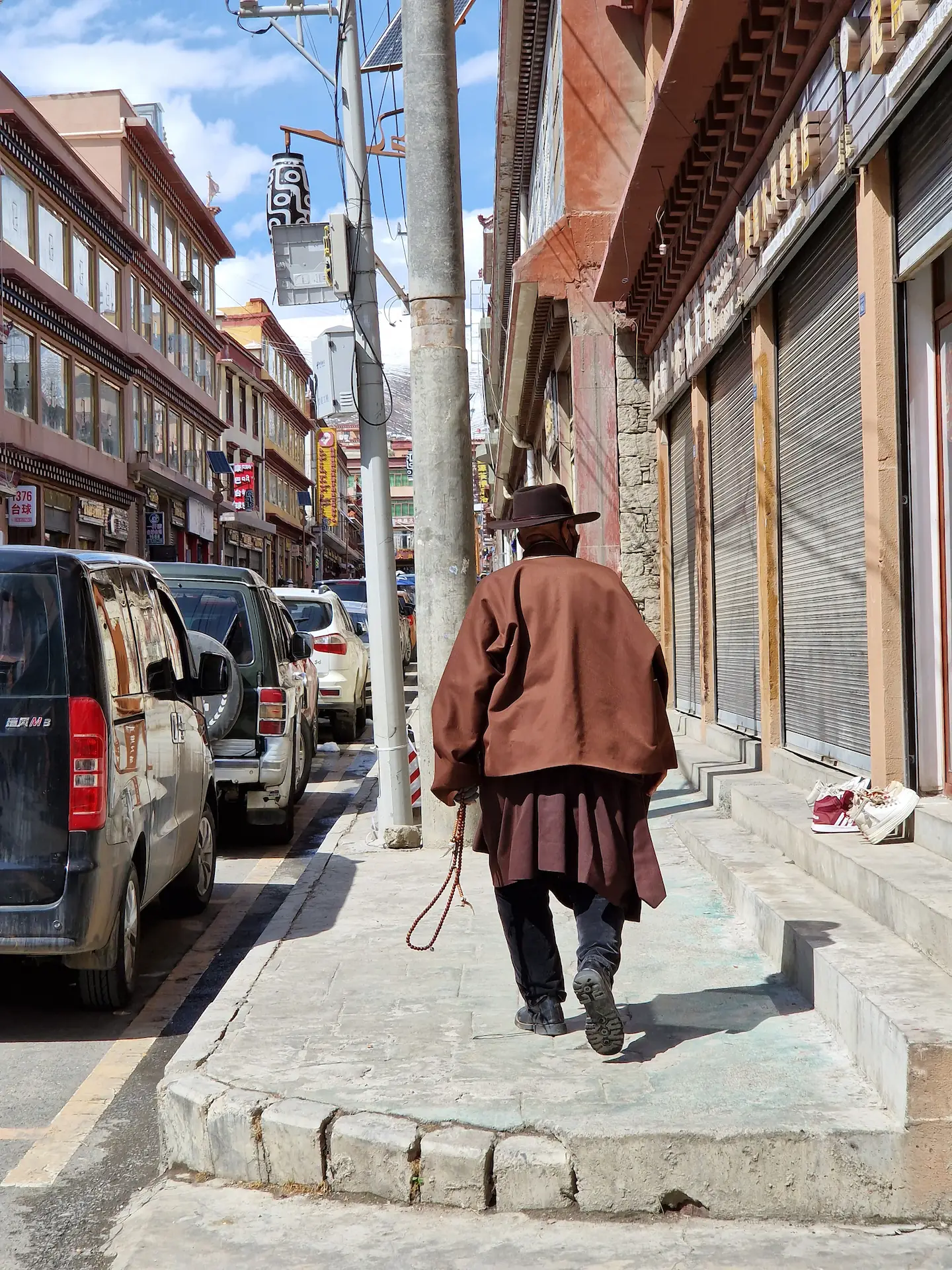 Un tibétain de dos, robe et pardessus bruns, chapeau de cowboy, chapelet trengwa à la main