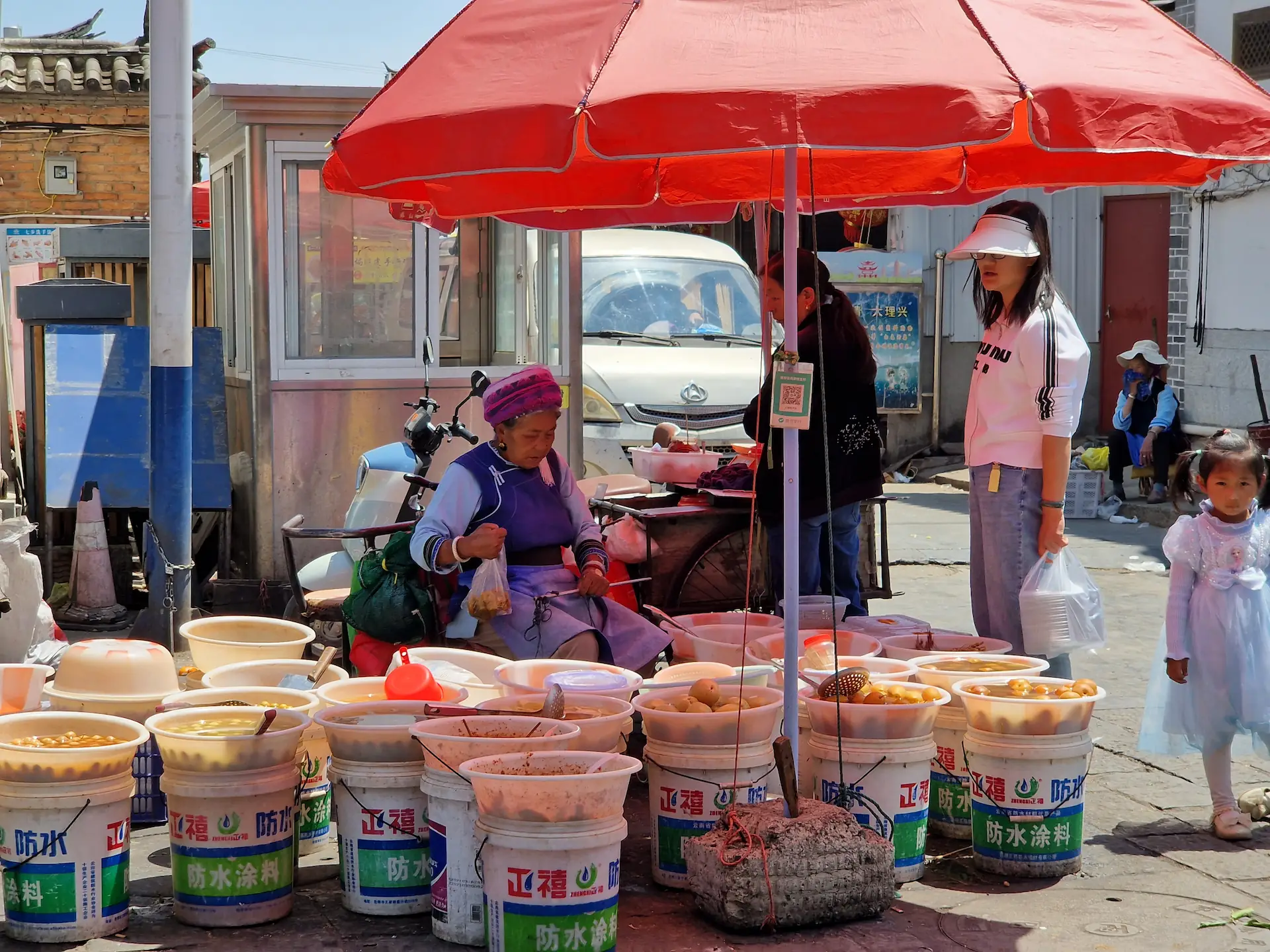 Une vielle vendeuse est assise au milieu de ses marchandises. Elle porte un habit bleu et une coiffe similaire à la première vendeuse.