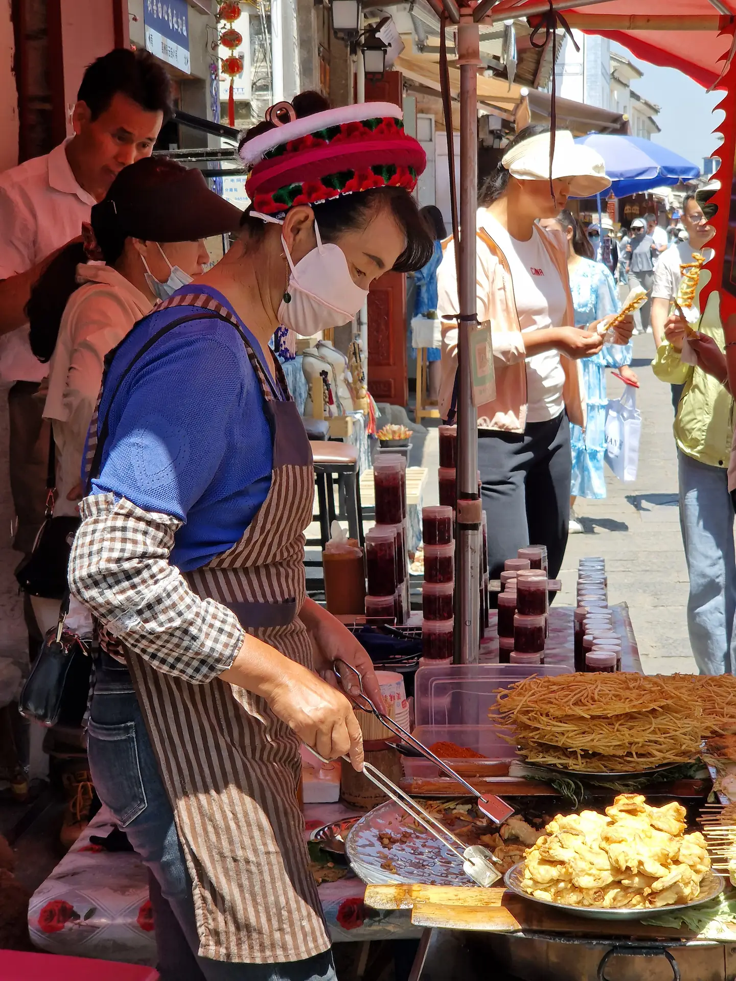 Une vendeuse au marché fait sauter des aliments dans une poêle. Elle porte une coiffe colorée qui retient ses cheveux.