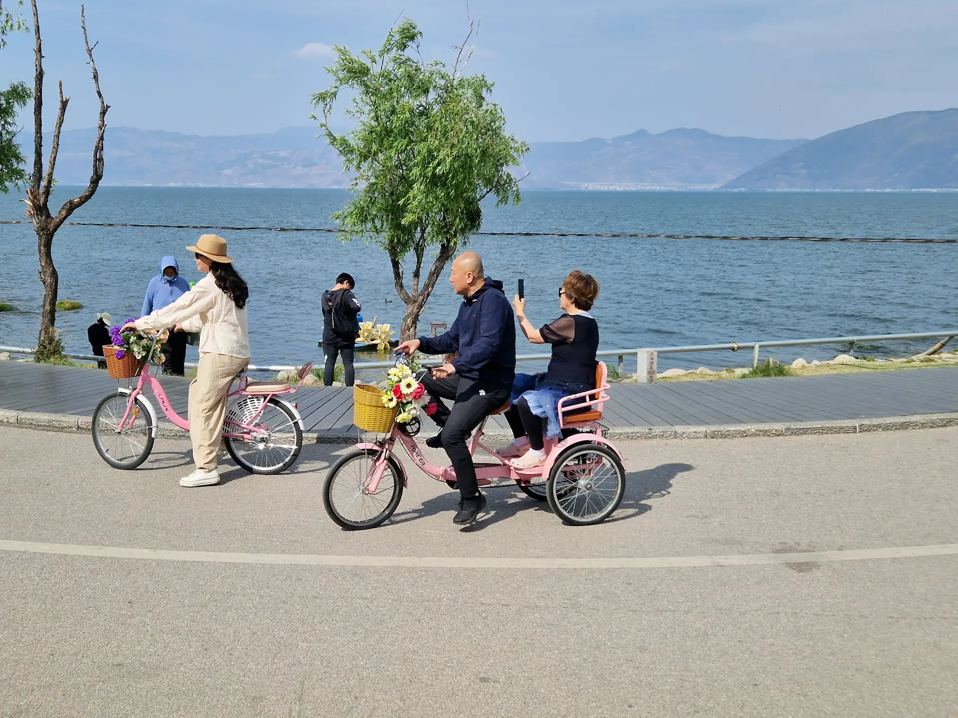 Un monsieur balade sa femme sur un tricycle rose avec panier à fleurs. La femme en profite pour prendre des photos du lac.