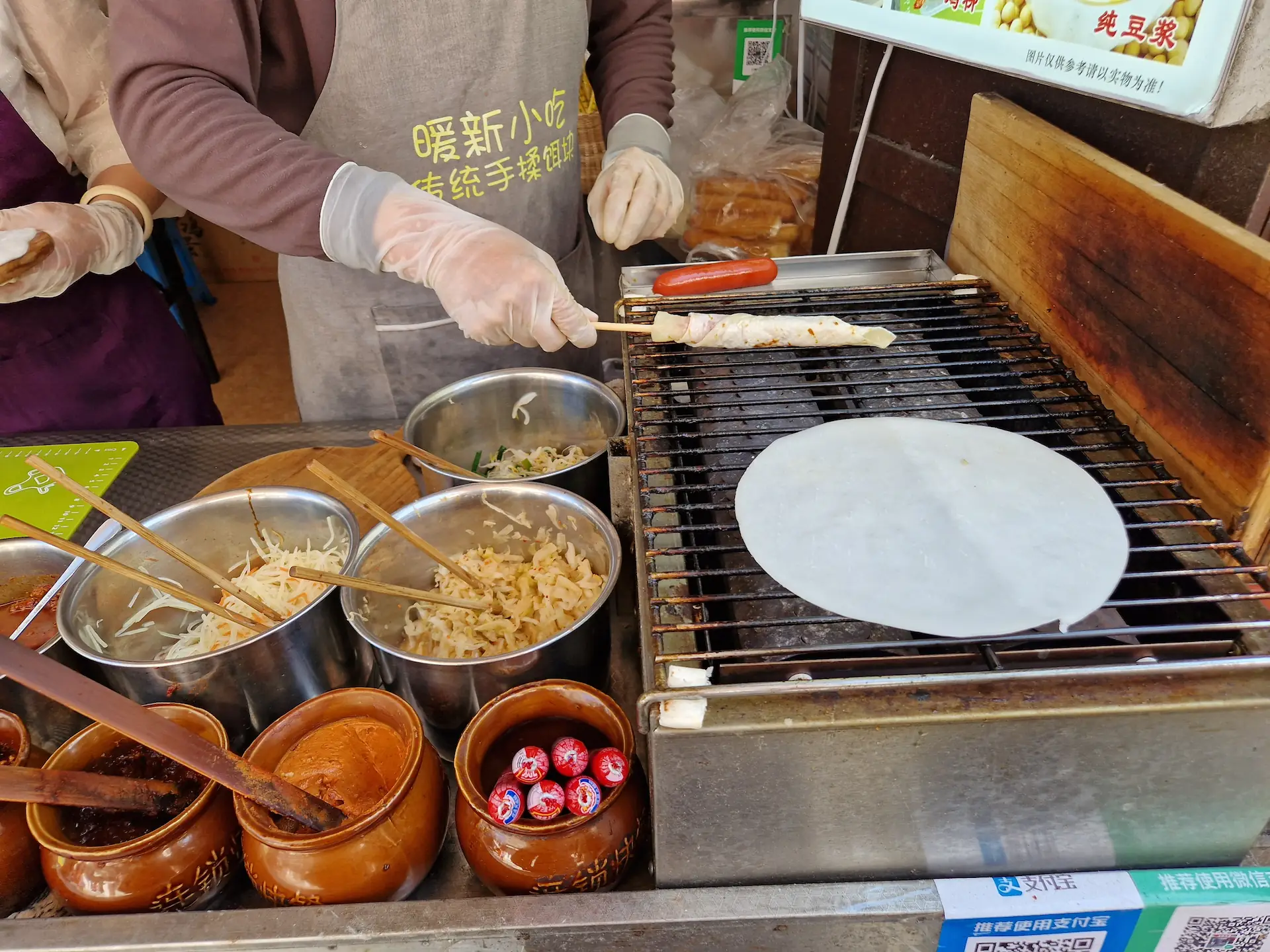 Sur une grille de BBQ dans la rue, un vendeur de street food nous prépare notre commande. Il enroule la crêpe au fromage autour d'un bâton alors qu'une galette de riz grille sur le côté.