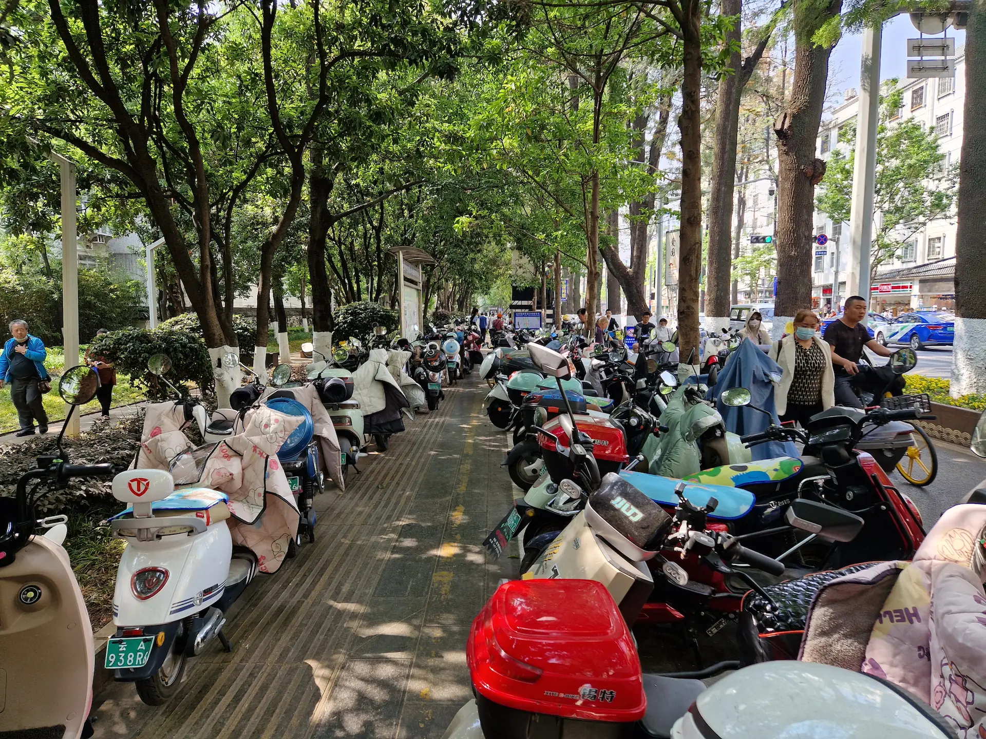 Des motos et scooters sont parquées le long d'un trottoir. On voit quelques piétons forcés de marcher sur la route.