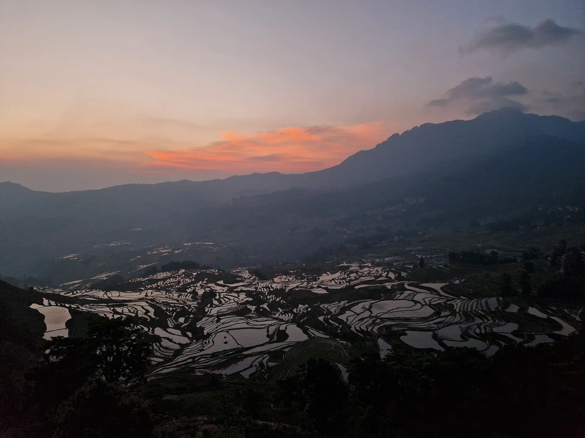 Le soleil va bientôt se lever sur les rizières de Duoyishu. Le ciel est encore endormi, les nuages sont roses-orangés. Les bassins à quelques centaines de mètres plus bas sont comme des miroirs sombres,