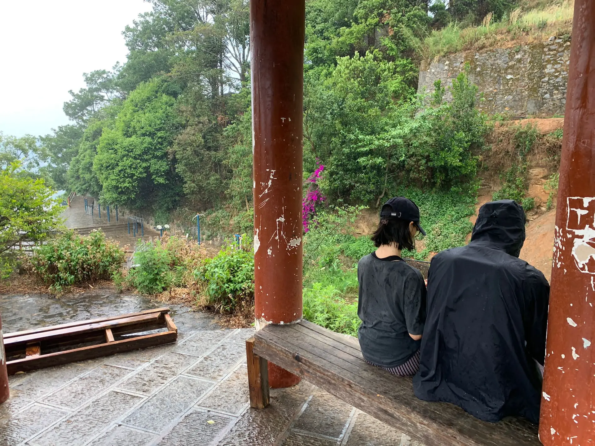 Clara et Robin, assis sur un banc sous le pavillon. La pluie tombe autour d'eux.